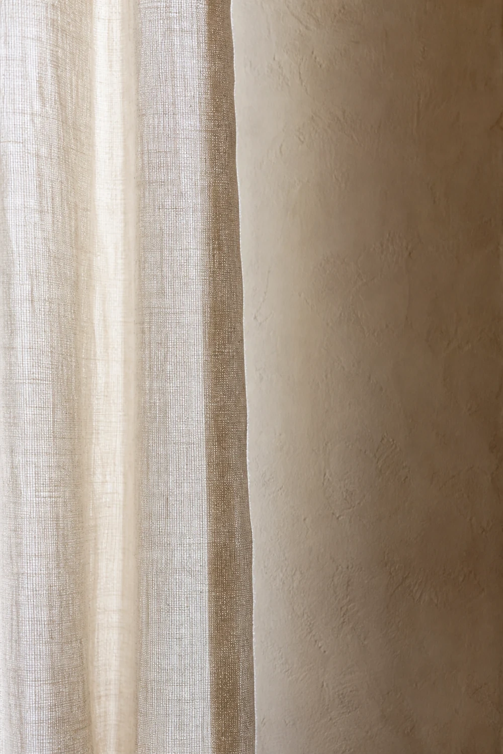 Sheer natural linen curtain fabric meets a textured warm plaster wall in soft diffused daylight.